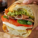 Handheld artisan prosciutto caprese sandwich on a sourdough baguette featuring buffalo mozzarella, heirloom tomatoes, fresh basil, and basil pesto, wrapped in brown butcher paper."
