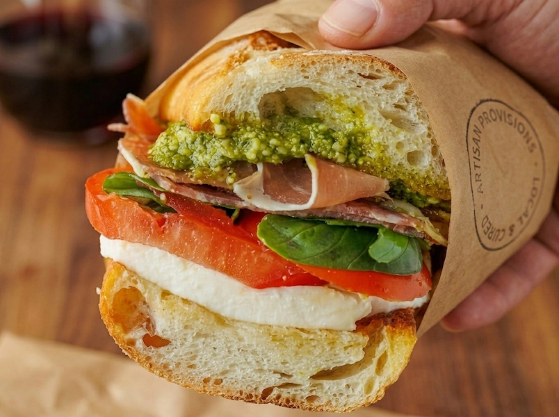 Handheld artisan prosciutto caprese sandwich on a sourdough baguette featuring buffalo mozzarella, heirloom tomatoes, fresh basil, and basil pesto, wrapped in brown butcher paper."