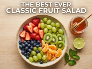 Top-down view of a colorful classic fruit salad bowl with strawberries, kiwi, grapes, and blueberries, alongside honey, lime, and mint for the dressing. Text reads The Best Ever Classic Fruit Salad.