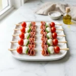 Mini antipasto skewers on a white platter, featuring cherry tomatoes, mozzarella balls, fresh basil, and folded salami slices, arranged neatly.