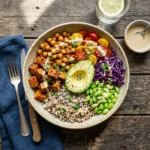 A vibrant vegetarian Buddha bowl featuring quinoa, roasted sweet potatoes, spiced chickpeas, avocado, edamame, cherry tomatoes, and red cabbage, drizzled with creamy tahini dressing.